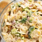 A bowl of broccoli florets, bacon bits, and shredded cheddar cheese mix in with tender bow tie pasta all coated in sweet homemade dressing.