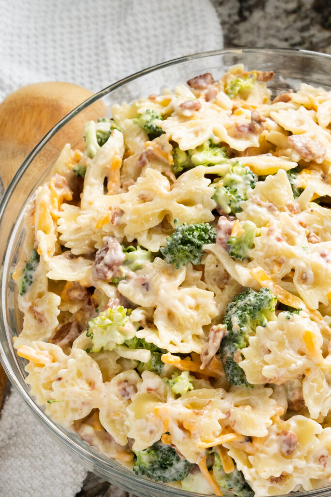 A bowl of broccoli florets, bacon bits, and shredded cheddar cheese mix in with tender bow tie pasta all coated in sweet homemade dressing.