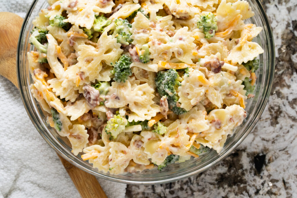 A bowl of broccoli florets, bacon bits, and shredded cheddar cheese mix in with tender bow tie pasta all coated in sweet homemade dressing.