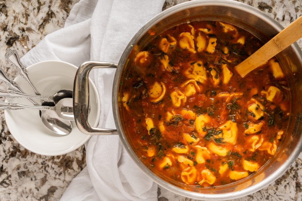 Overview of spinach tortellini soup in a large stock pot with a bowl and spoons to the left.