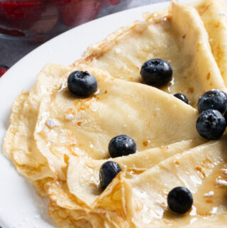 Close up of crepes folded drizzled in syrup and sprinkled with powdered sugar and blueberries ready to be served.