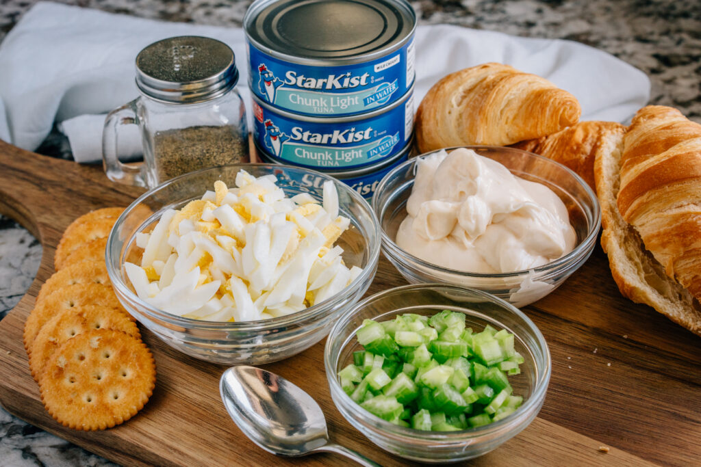 Canned Tuna, Diced hard-boiled eggs, chopped celery, Miracle Whip, and pepper all laid out on a cutting board with croissants and butter crackers.
