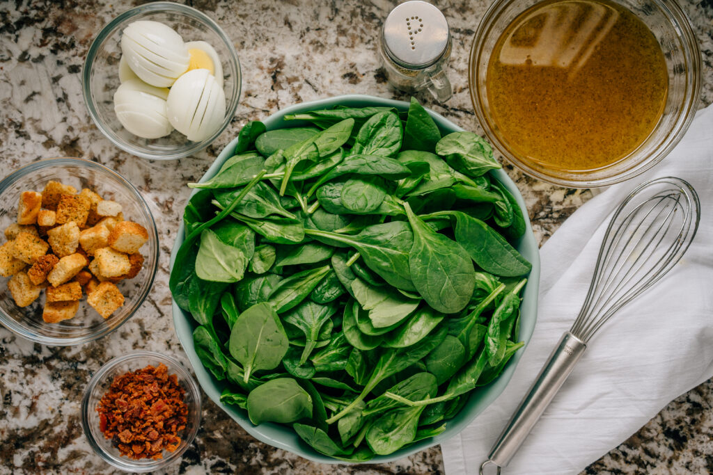 All ingredients for a egg and bacon spinach salad in seperate bowls.