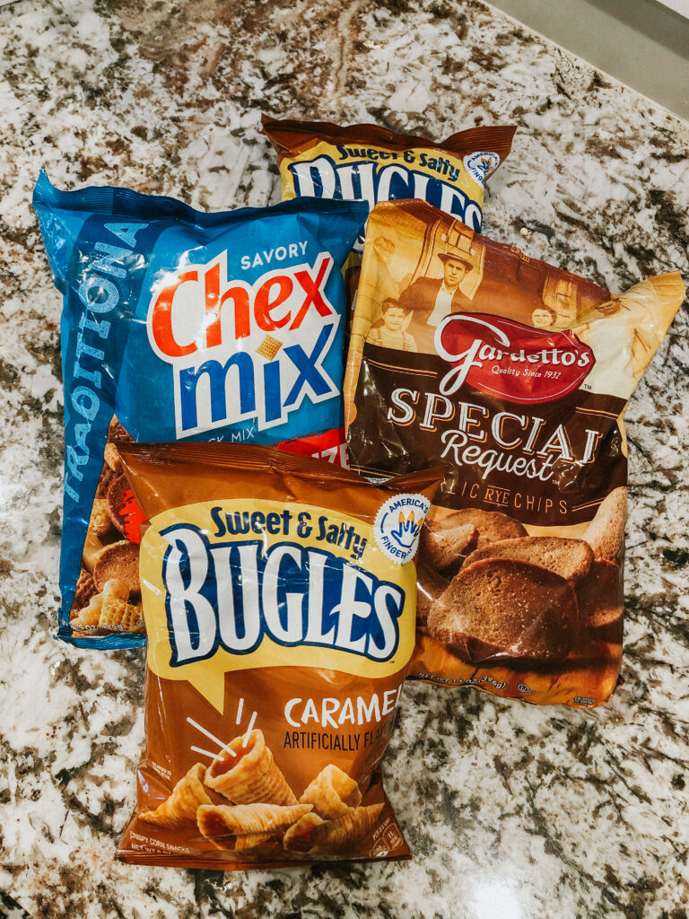 A bag of family sized chex mix, rye chips, and two bags of caramel bugles.