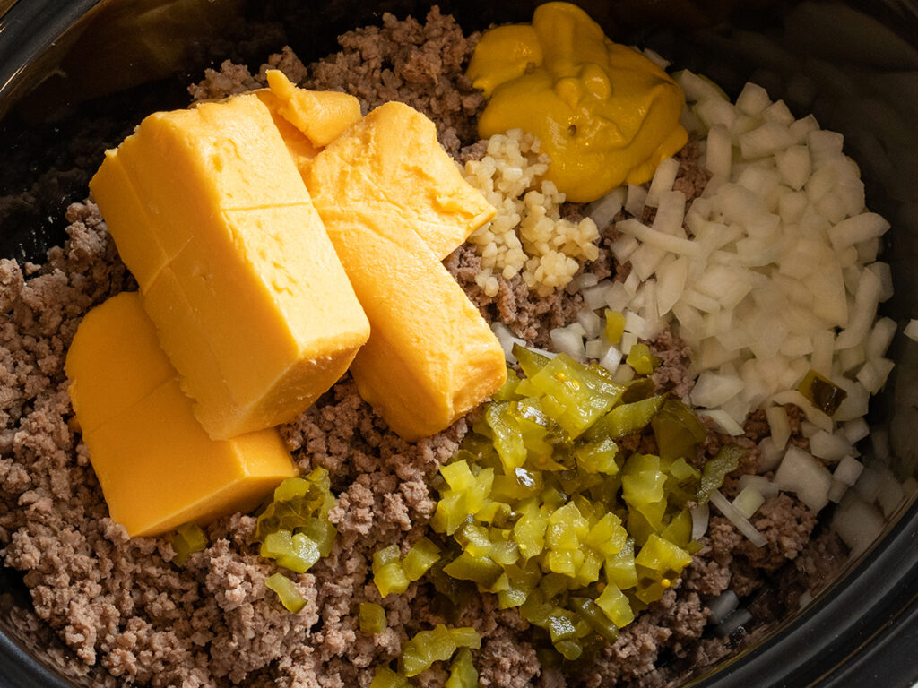 Chopped onion and pickles, garlic, mustard, velveeta cheese and browned ground beef are sitting in a crockpot ready to be stir and cook to make crockpot cheeseburgers!