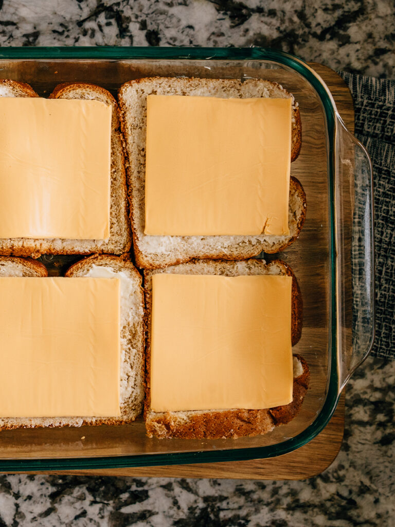 Image of buttered bread(buttered side up) with slice cheese on top in a baking dish. Six of these breads are the bottom layer of this casserole.