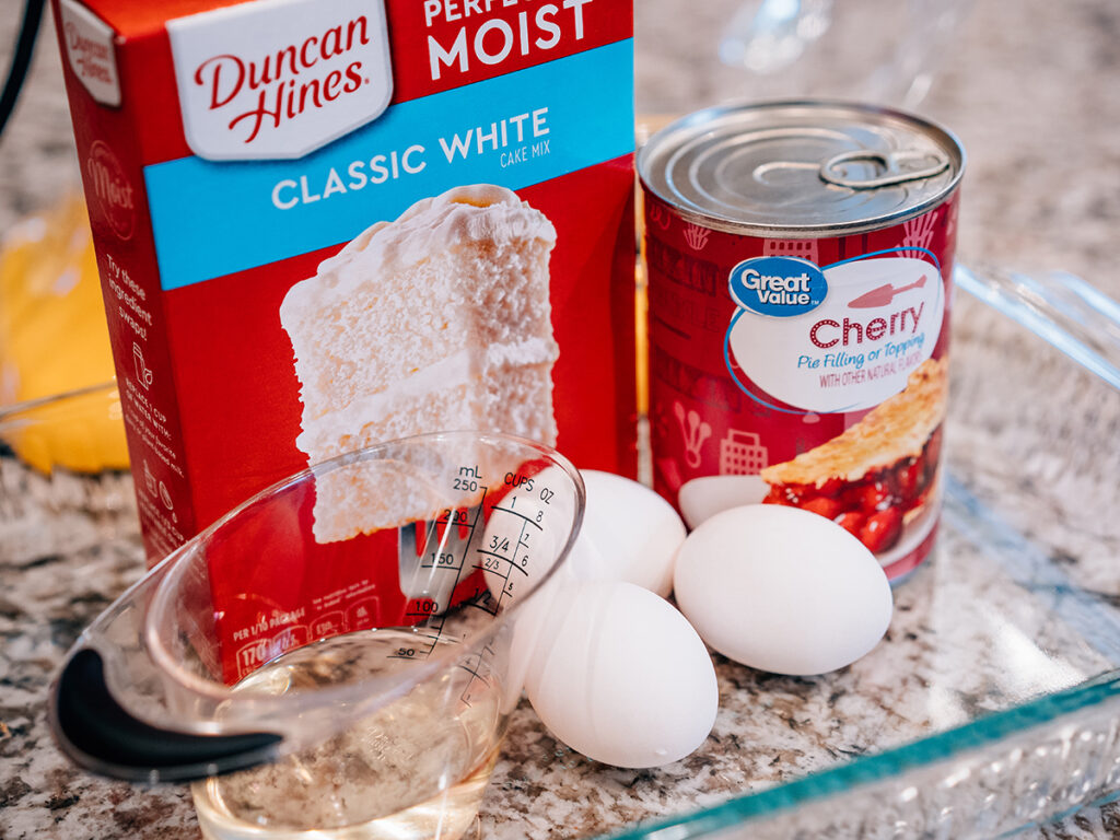 A box of white cake mix, a can of cherry pie filling, eggs and water all in a pyrex dish. These are all the ingredients of this delicious cherry bruch cake.