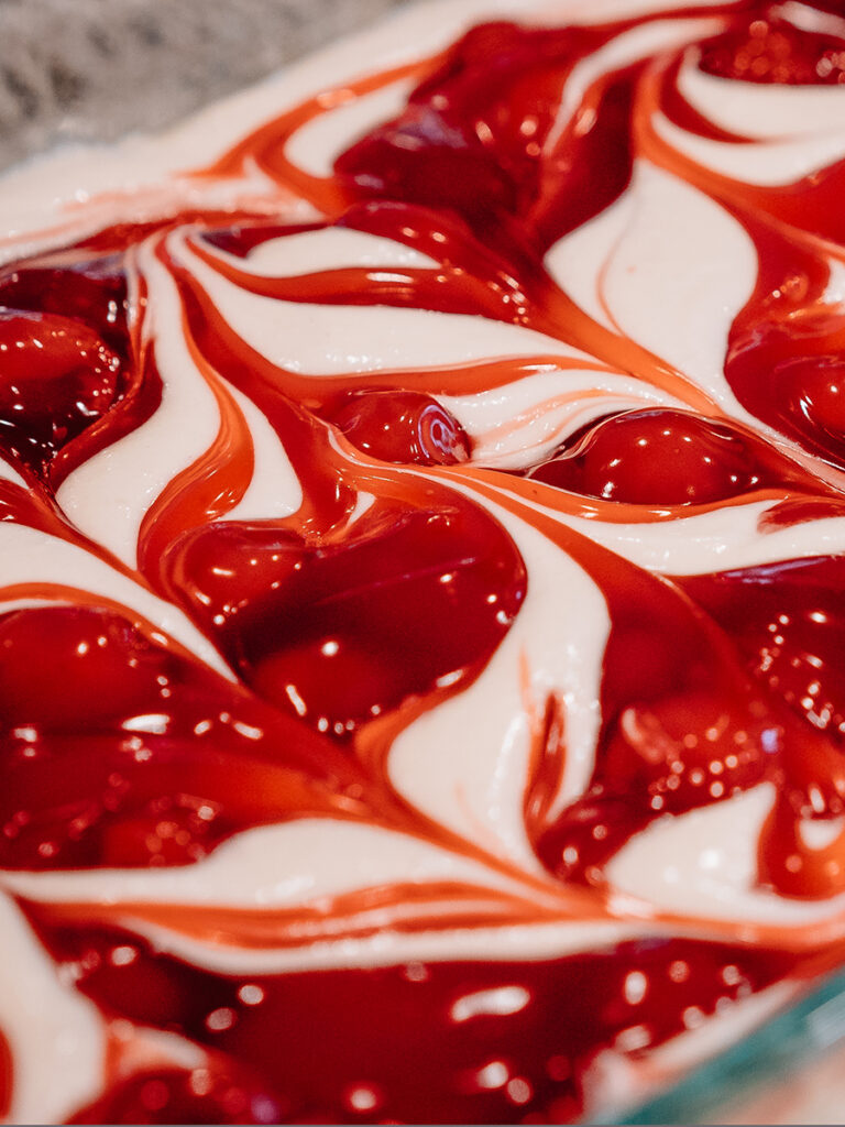 A close-up photo of cherry pie filling swirled into white cake batter.