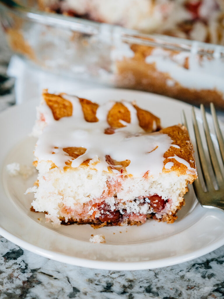 A plated piece of delicious cherry brunch cake topped with a simple powdered sugar, milk, and vanilla extract glaze.