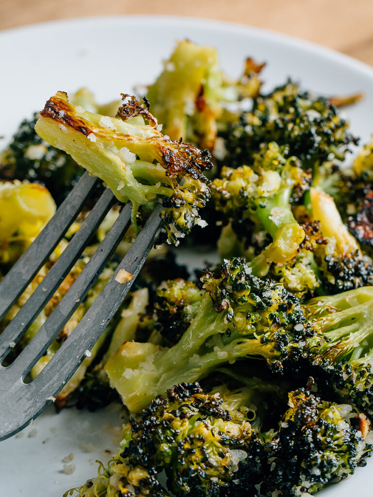 A piece of tender roasted broccoli with parmesan speared by a fork, warm, flavorful, and ready to eat!