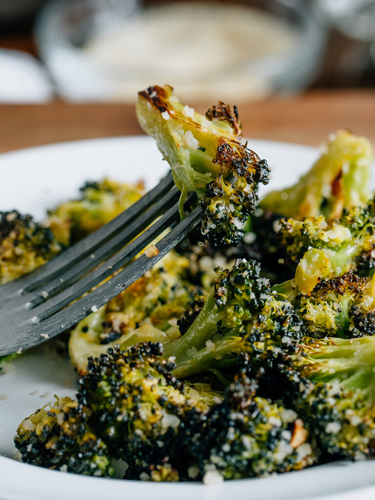 A piece of tender roasted broccoli with parmesan speared by a fork with a serving more in the background: warm and flavorful broccoli. Ready to eat!