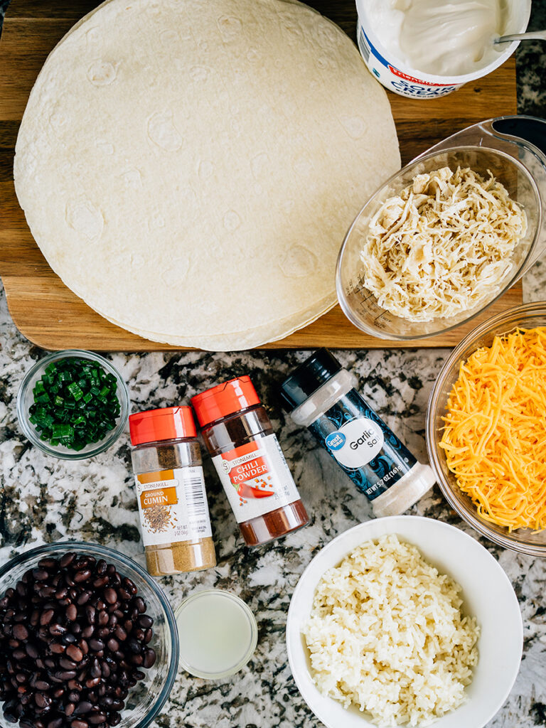 The ingredients of the crispy southwest chicken wrap all laid out on the counter: Rice, shredded chicken, black beans, flour tortillas, lemon juice, shredded cheese, sour cream, green onions, and spices.
