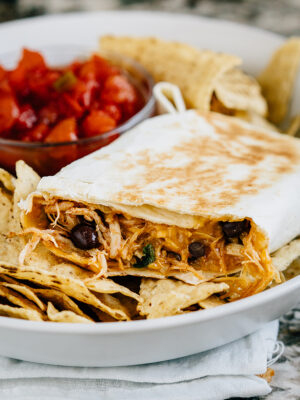 A crispy southwest chicken wrap with a couple of bites taken out of it laying on a bed of tortilla chips with salsa in the background.