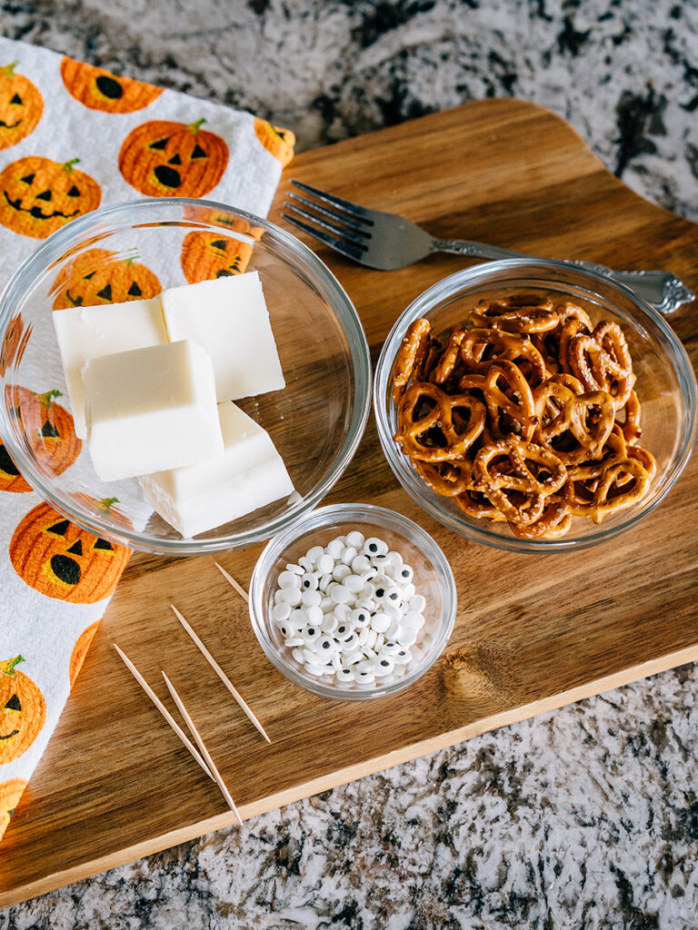 Vanilla almond bark, twisted pretzels, and candy eyes in prep bowls ready to make Halloween ghost faces.