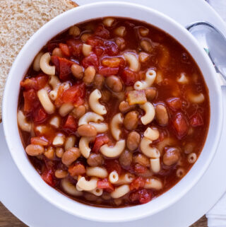 Baked bean and macaroni soup in a white bowl with a buttered bread on the side.