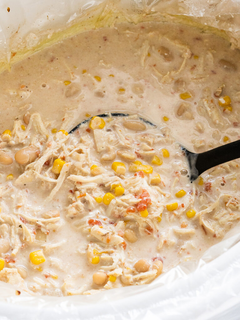 A crock pot filled with bubbling white chicken chili: shredded chicken, great northern beans, corn, Rotel, onion and spices in a creamy broth garnished with corn chips on the side.