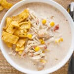A bowl of shredded chicken, great northern beans, corn, Rotel, onion and spices in a creamy broth garnished with corn chips on the side.