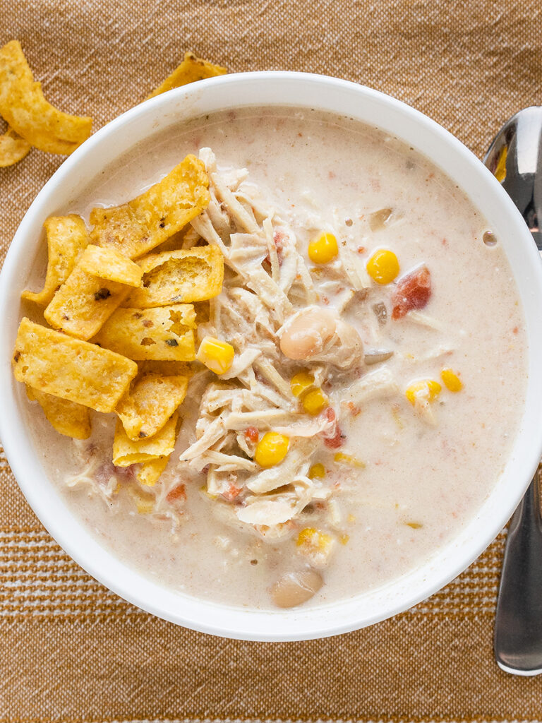 A bowl of shredded chicken, great northern beans, corn, Rotel, onion and spices in a creamy broth garnished with corn chips on the side.