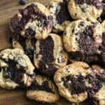 Pile of delicious Brookie Cookies on a cutting board with chocolate chips scattered in front.