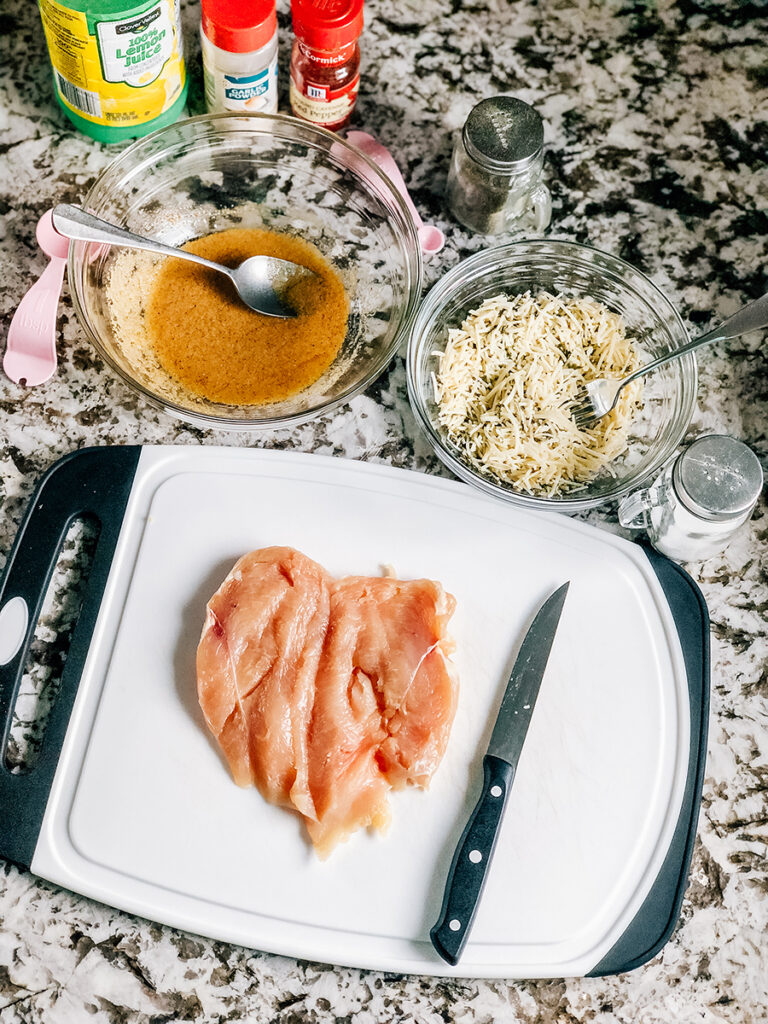 A raw butterflied chicken breast on a cutting board, a bowl of garlic marinade, and a bowl of parmesan cheese topping.