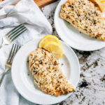 A plated and sliced baked garlic parmesan chicken breast with lemon slices on the side.
