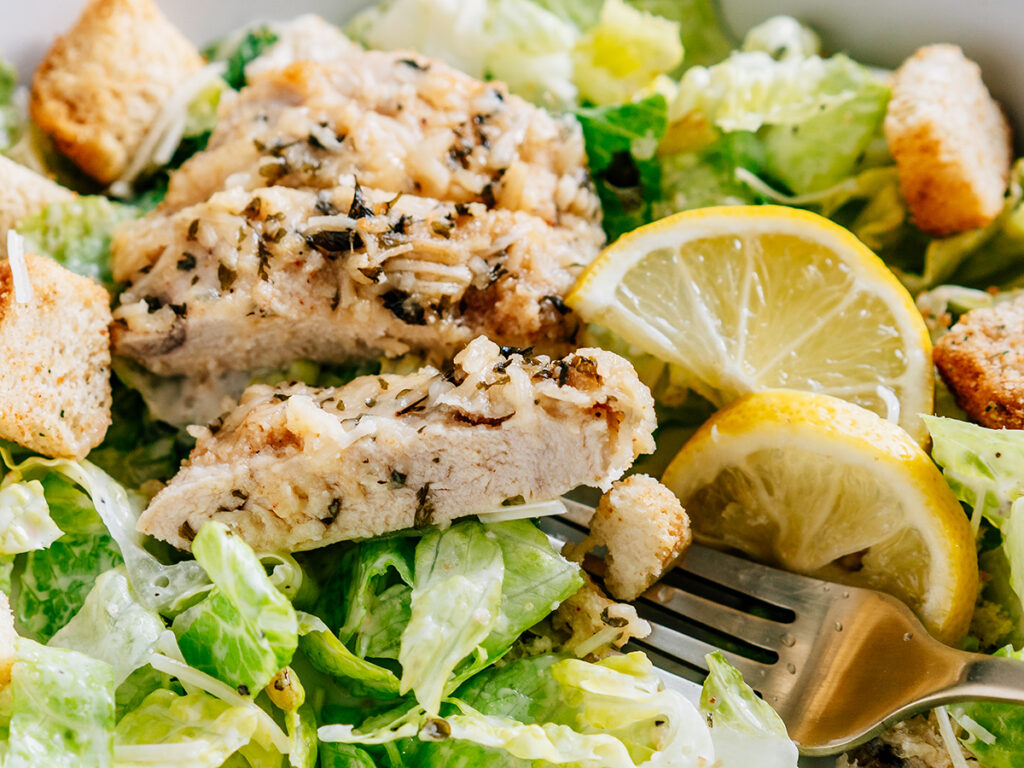 Close up of a bowl of fresh romaine lettuce covered in cesar salad dressing topped with crunchy cortons, a sprinkle of parmesan cheese and a tender and juicy piece of baked garlic parmesan chicken.