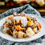 A serving of tender potatoes and carrots with browned ground hamburger baked in butter and onions sprinkled with shredded cheddar cheese and topped with a dollop of sour cream piled high on a plate ready to eat!