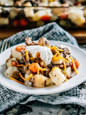 A serving of tender potatoes and carrots with browned ground hamburger baked in butter and onions sprinkled with shredded cheddar cheese and topped with a dollop of sour cream piled high on a plate ready to eat!