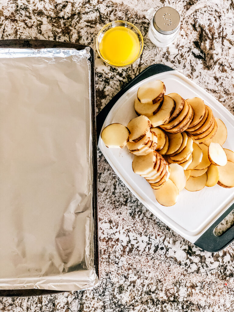 Baking sheet lined with foil next to a cutting board with a pile of freshly cut Russet potato slices, melted butter, and a salt shaker.
