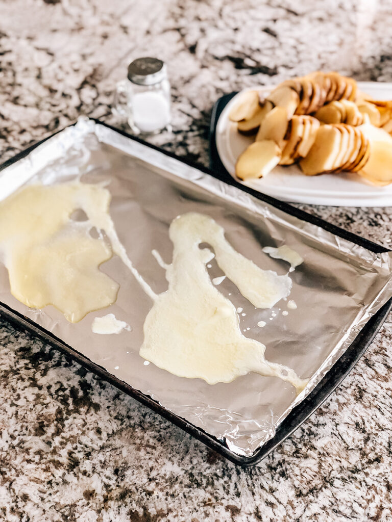 Baking pan lined with foil and drizzled with melted butter with potato slices in the background on a cutting board.