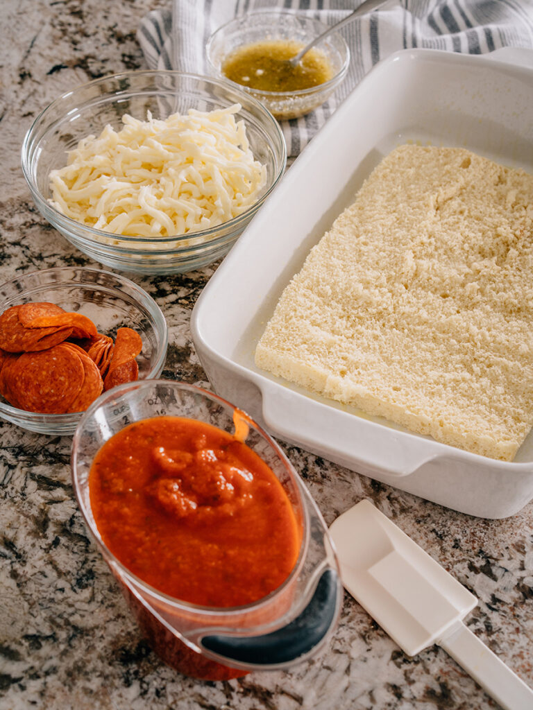 A picture of all the ingredients that make pizza sliders: shredded mozzarella cheese, pizza sauce, pepperoni, sweet Hawaiian rolls, and a yummy butter sauce.
