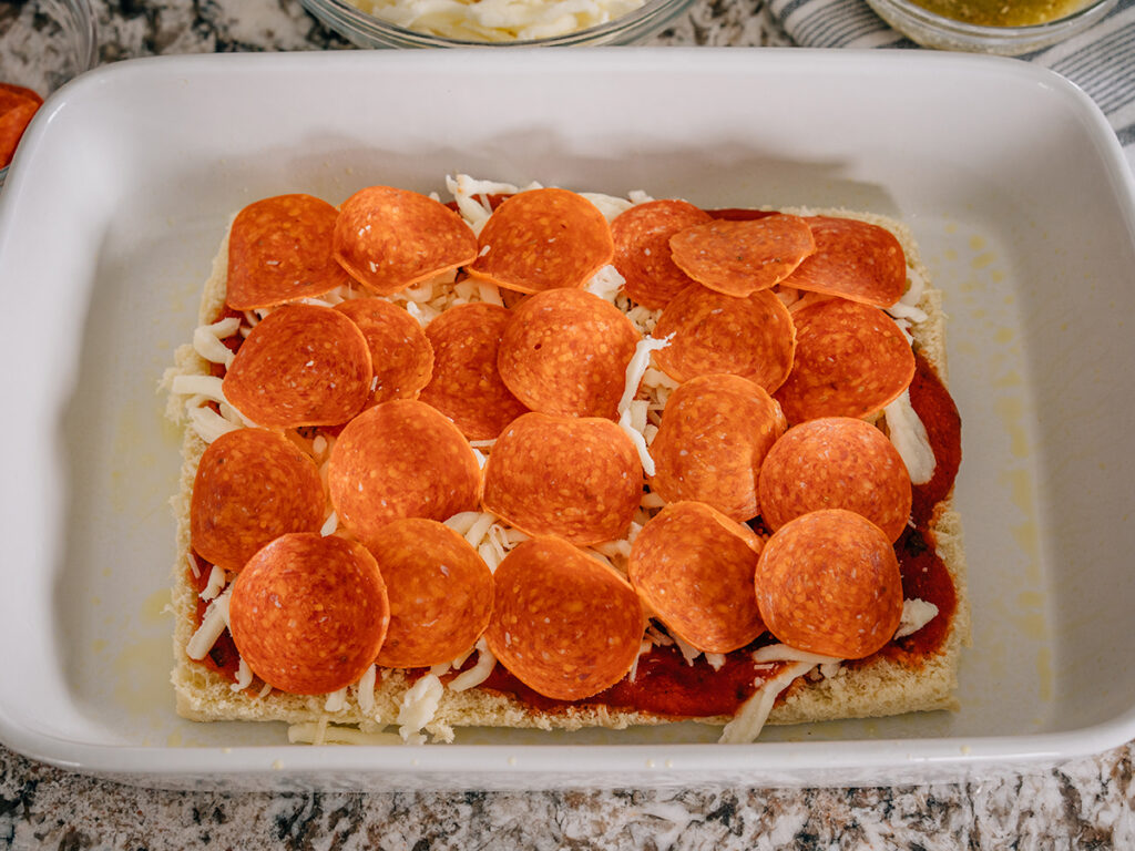 A slab of Hawaiian rolls in a baking dish with spaghetti sauce, shredded mozzarella cheese, and pepperoni slices layered on top.