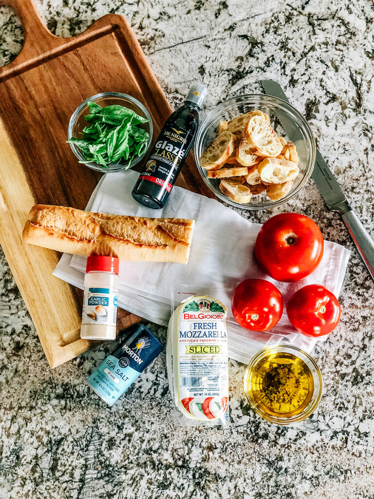 Ingredients for deconstructed bruschetta: fresh mozzarella slices, tomatoes, basil leaves, and a french baguette loaf.