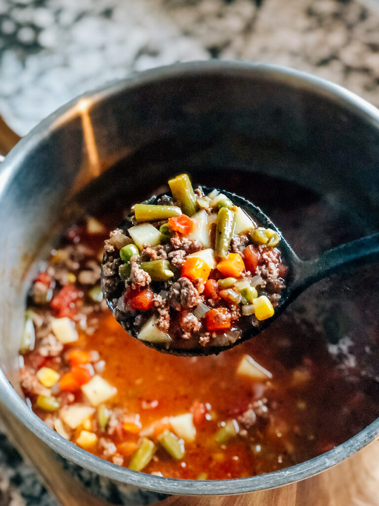 A ladle of hamburger soup with frozen vegetables hot and ready to be served.