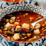 A spoon full of hamburger soup that has corn, green beans, carrots, potatoes, onions and other seasonings.