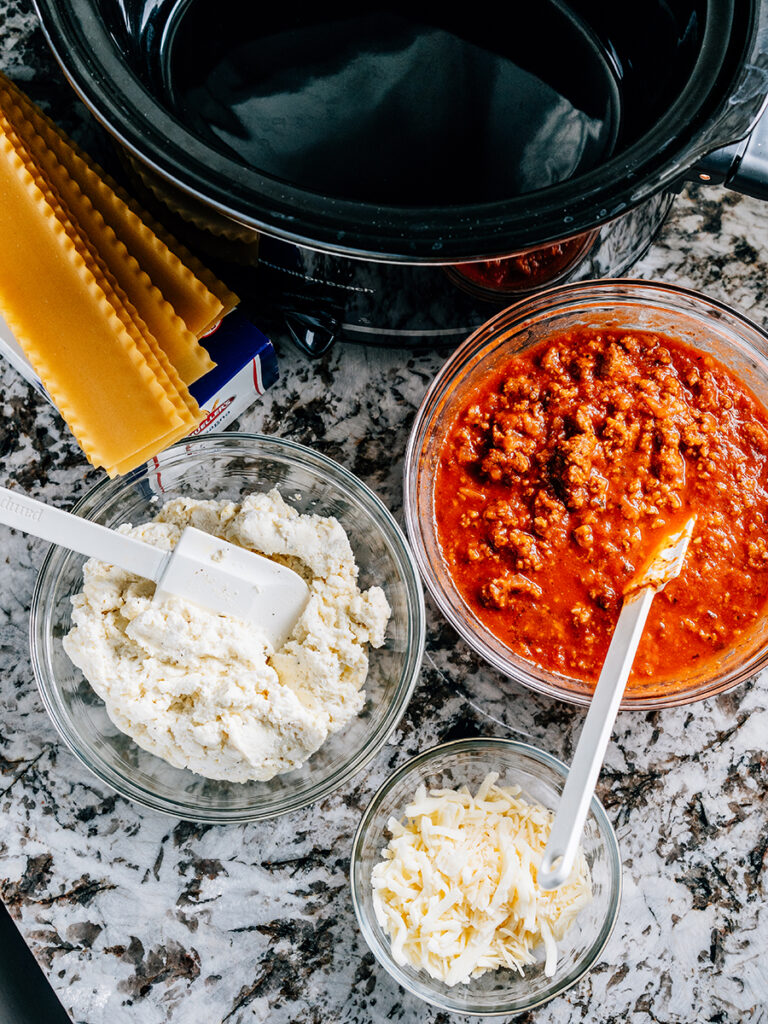 Lasagna noodles, meat sauce, cheese mixture and cheese topping all in bowls waiting to be assembled into the easiest crockpot lasagna.