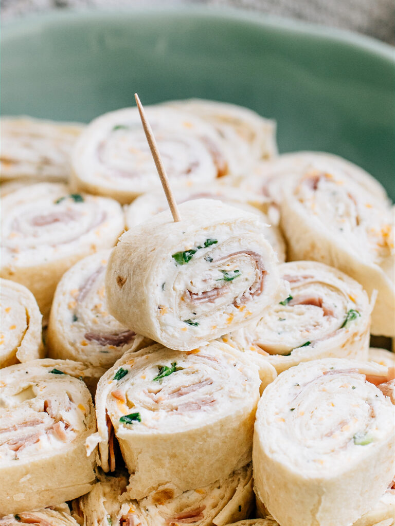 A single ham and cheese roll up with a toothpick in it on top of other roll ups waiting to be served.