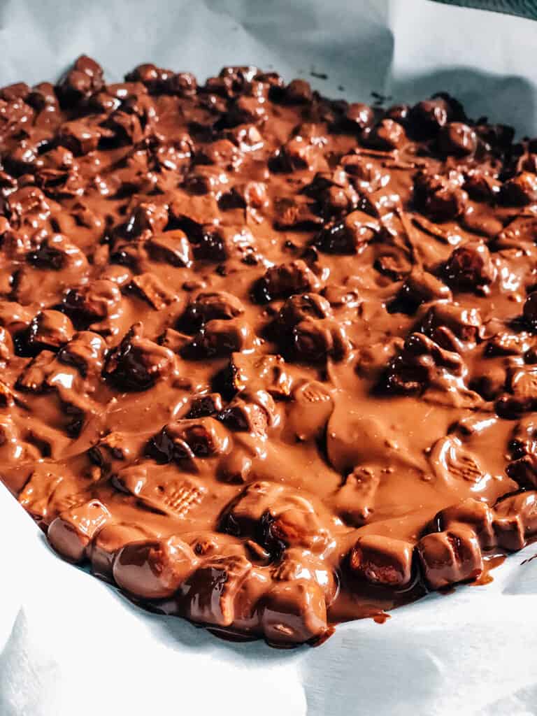 The thin layer of chocolate with golden grahams and mini marshmallows spread thin on parchment paper.