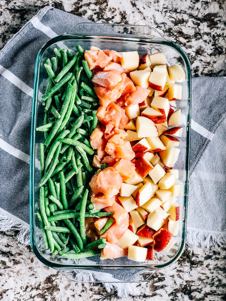 A 9x13 inch baking pan with rows of frozen green beans, raw chicken breasts cubes and red potatoes.