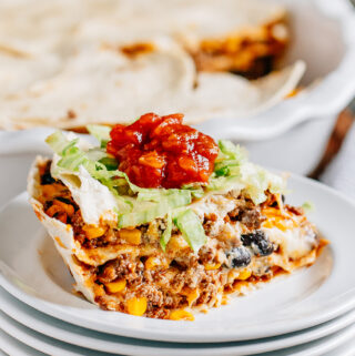 A slice of taco pie showing off the layers of tortillas, taco meat with corn and black beans, sour cream, and cheese then topped with a garnish of shredded iceberg lettuce and a dollop of salsa. SO Good!