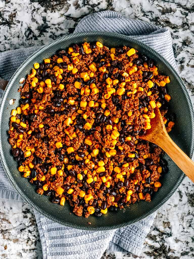 The skillet with the browned ground meat made into taco meat mixed with the canned corn and drained and rinse black beans.