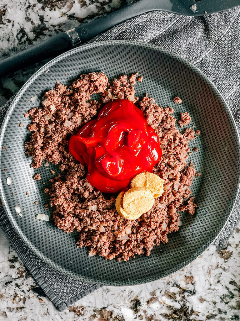 A skillet with browned ground beef, ketchup and light brown sugar in it.