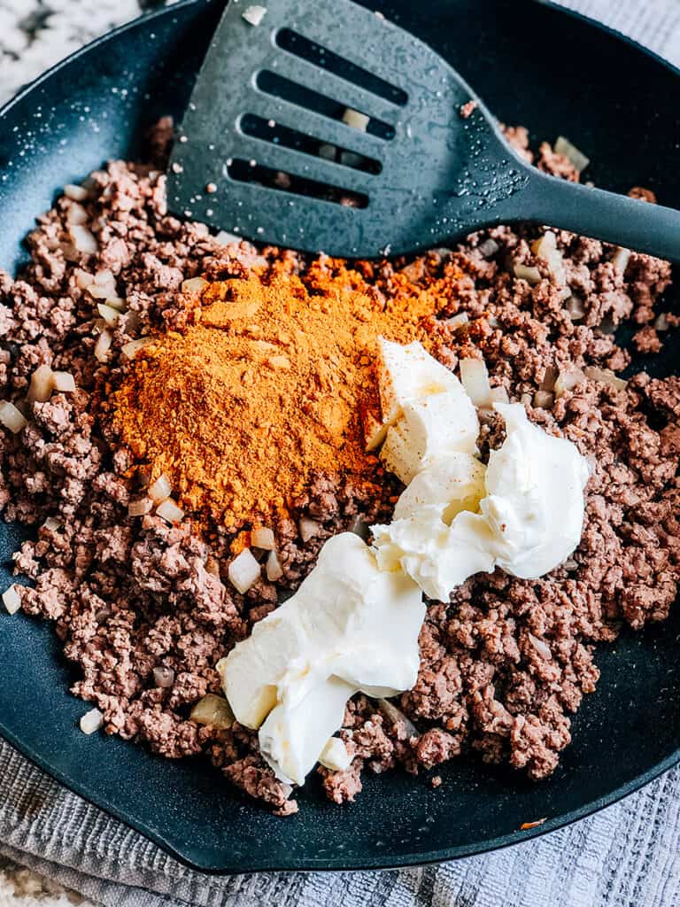 Browned ground beef with chopped onion in a skillet with taco seasoning and softened cream cheese on top ready to be mixed.