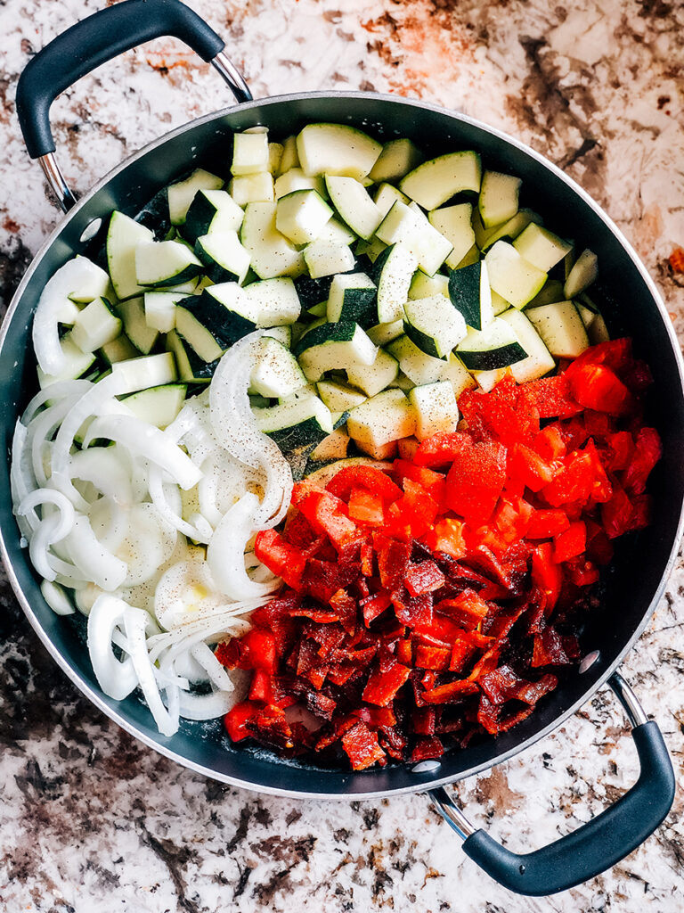 A skillet loaded full of diced tomatoes and zucchini. crumbled bacon, and sliced onions seasoned with salt and pepper.
