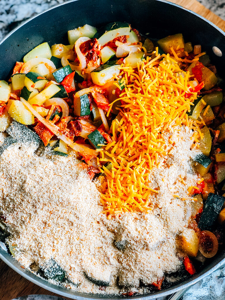 A skillet full of cooked garden vegetables with shredded cheddar cheese, bread crumbs, and grated parmesan cheese and soy sauce added and ready to be mixed.