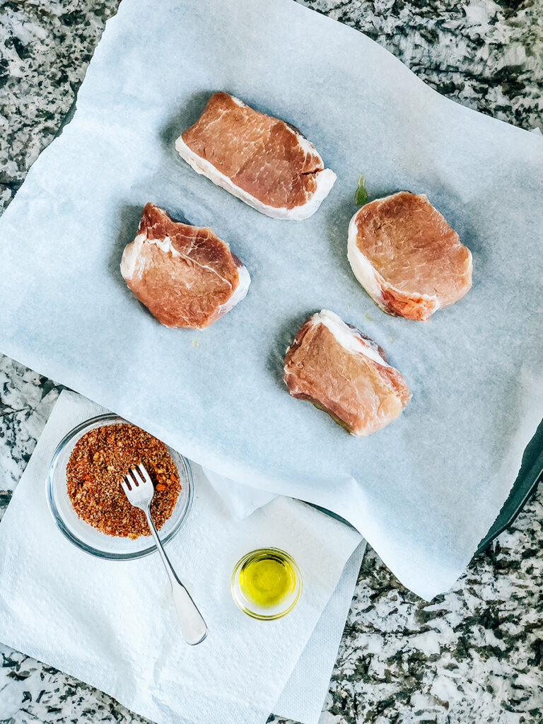 An overview image showing four raw pork chops on a baking sheet, a bowl of perfect pork chop spice rub, and some olive oil.