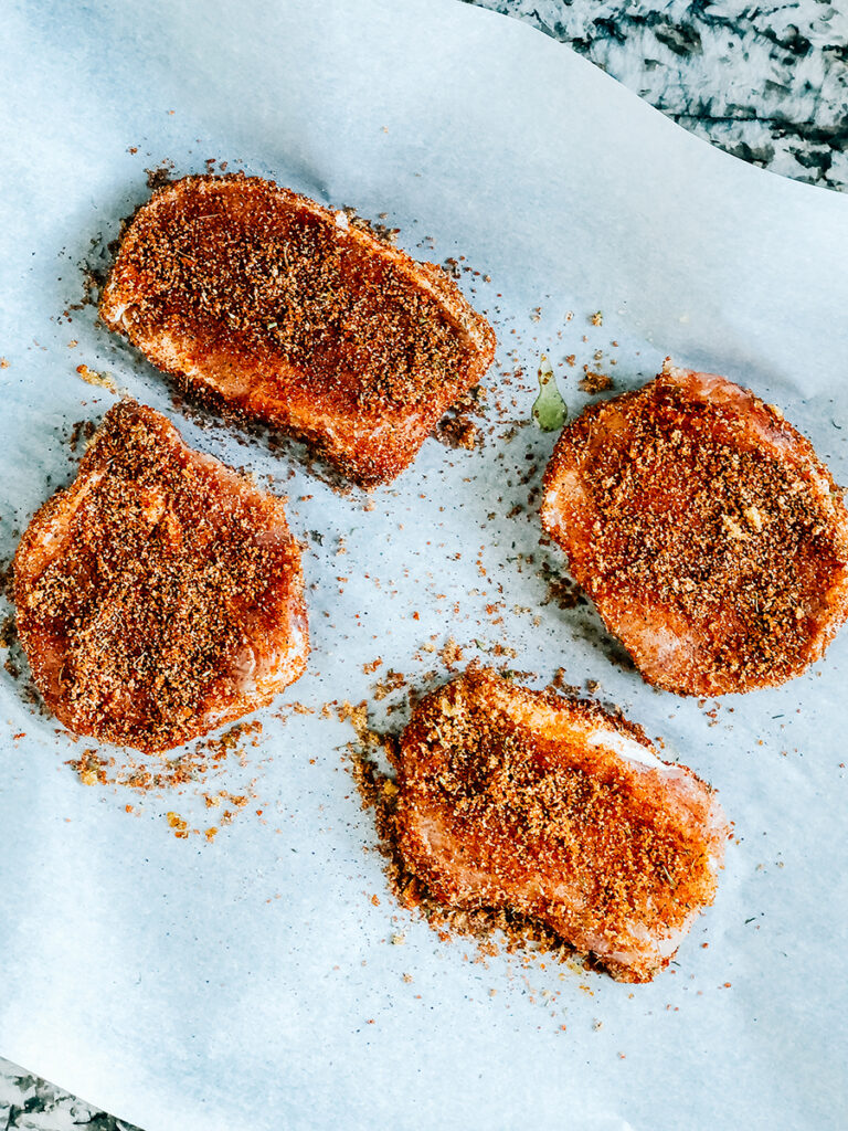 Four raw pork chops completely covered in a sweet spice rub ready to be baked.