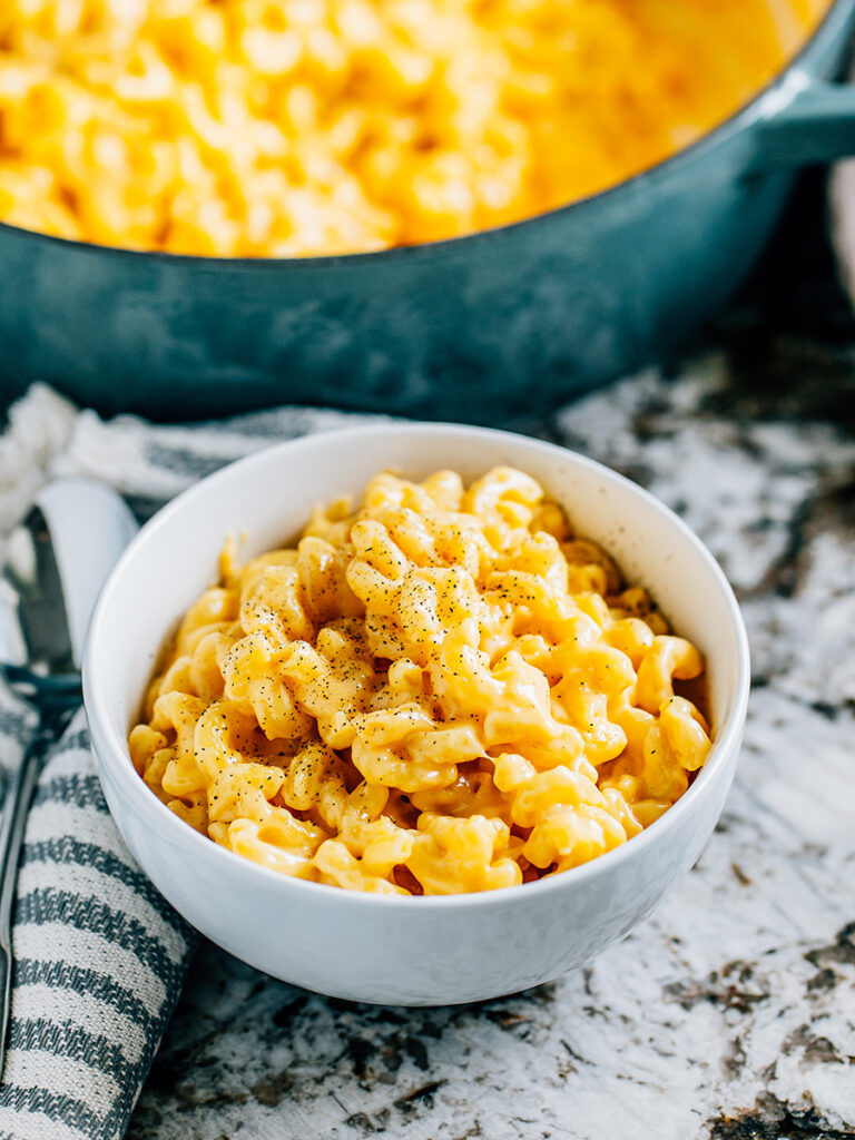 A finish bowl of creamy homemade mac and cheese with a sprinkle of black pepper on top in front of the skillet of mac and cheese.