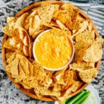 Slow cooker creamy buffalo chicken dip in a serving bowl surrounded by thick and sturdy tortilla chips with a plate of celery sticks close by and the slow cooker with extra dip in the corner.