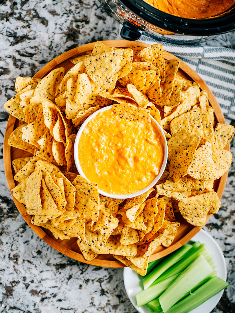 Slow cooker creamy buffalo chicken dip in a serving bowl surrounded by thick and sturdy tortilla chips with a plate of celery sticks close by and the slow cooker with extra dip in the corner.